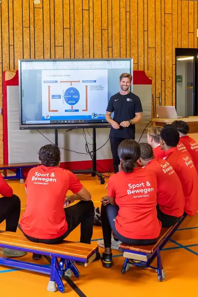 Groep studenten in rode shirts met 'Sport & Bewegen' luistert in de gymzaal naar een docent.