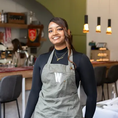 Een jonge vrouw met een schort staat lachend in een horecazaak. Op de achtergrond is een bar te zien met barkrukken, koffieapparaten en verlichting.