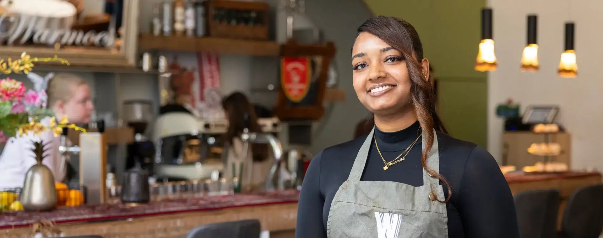 Een jonge vrouw met een schort staat lachend in een horecazaak. Op de achtergrond is een bar te zien met barkrukken, koffieapparaten en verlichting.