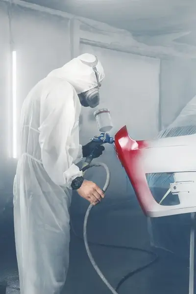 Student in overall verft auto-onderdeel met spuitbus in werkplaats.