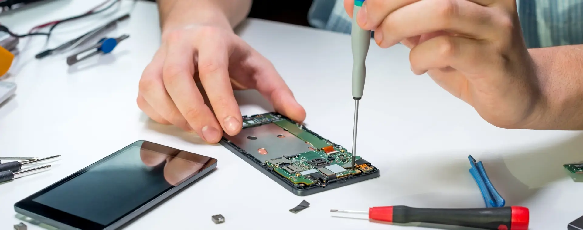 Close-up van handen die een smartphone repareren met gereedschap op een witte werktafel.