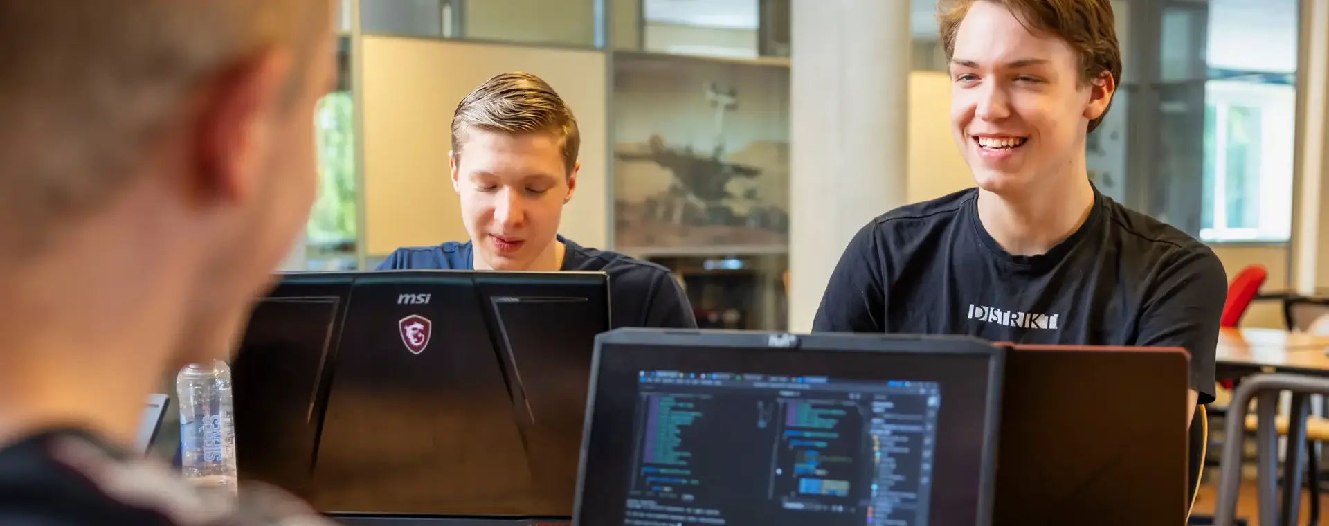 Drie studenten werken samen aan een programmeeropdracht. Ze zitten met hun laptops aan een tafel en overleggen enthousiast over hun code.