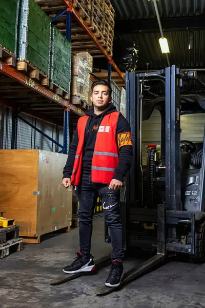 Jongen staat op vorken van zijn vorkheftruck in een magazijn.
