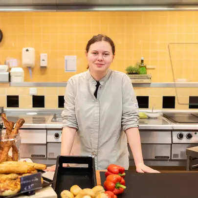 Een student in een keukenvest staat achter een toonbank met vers eten en kijkt zelfverzekerd in de camera. 