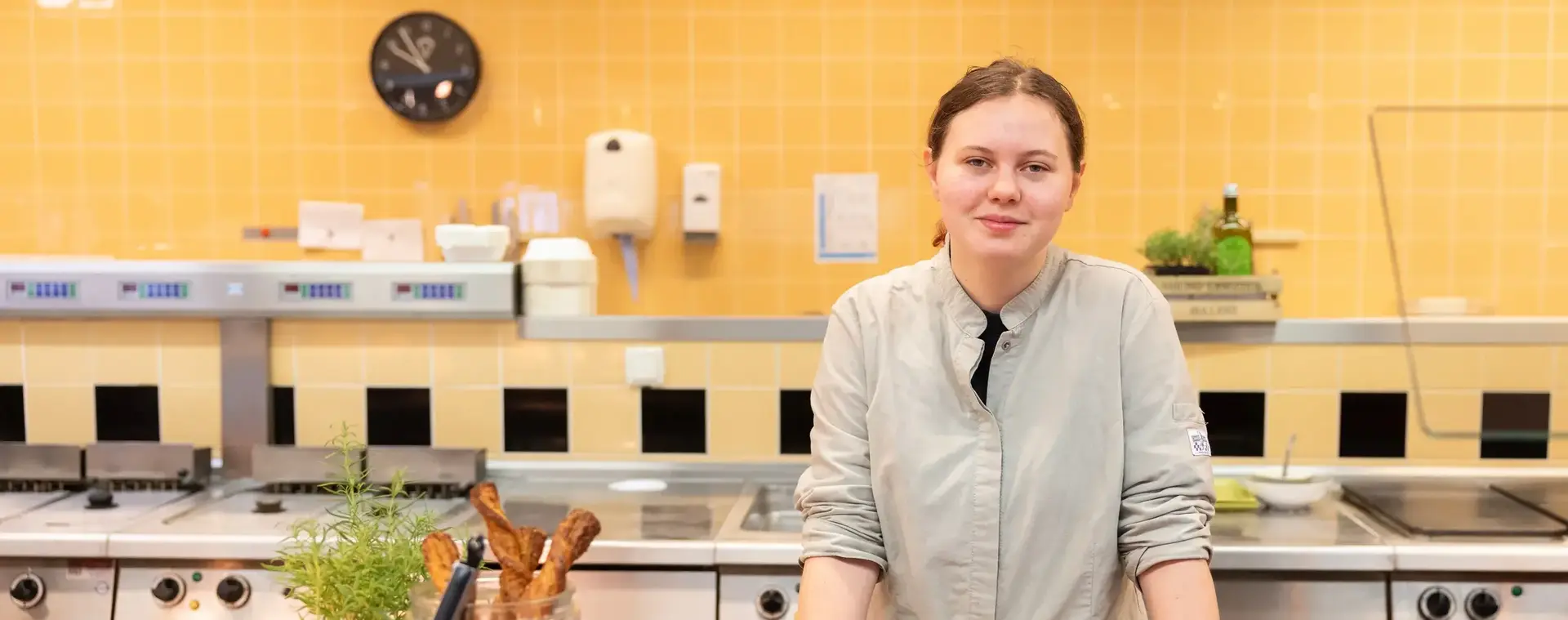 Een student in een keukenvest staat achter een toonbank met vers eten en kijkt zelfverzekerd in de camera.