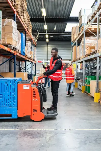 Student in rood hesje staat op een kleine vorkheftruck in een opslagloods.
