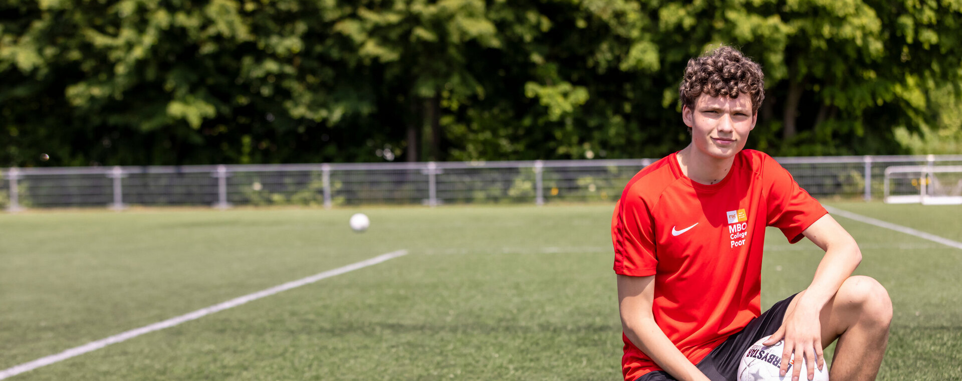 Een student in sportkleding knielt op een voetbalveld en houdt een bal vast. Op zijn shirt is het logo van MBO College Poort zichtbaar.