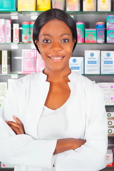 Apotheker in witte jas glimlacht voor een wand met medicijnen en doosjes in een apotheek.