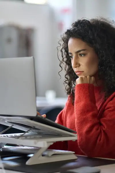 Student werkt geconcentreerd achter een laptop op kantoor. Ze draagt een rode trui en steunt met haar hand onder haar kin terwijl ze naar het scherm kijkt.