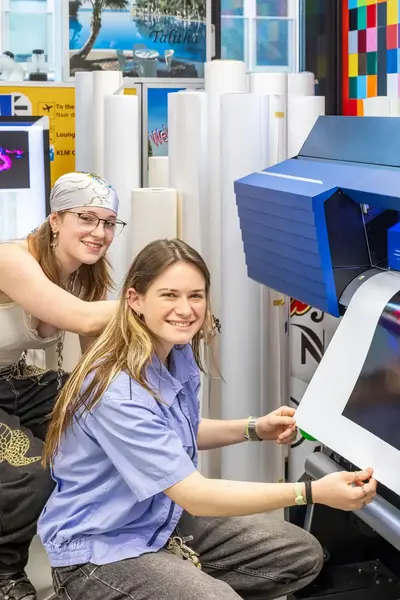 Twee studentes halen een grote glanzende sticker uit een printer.