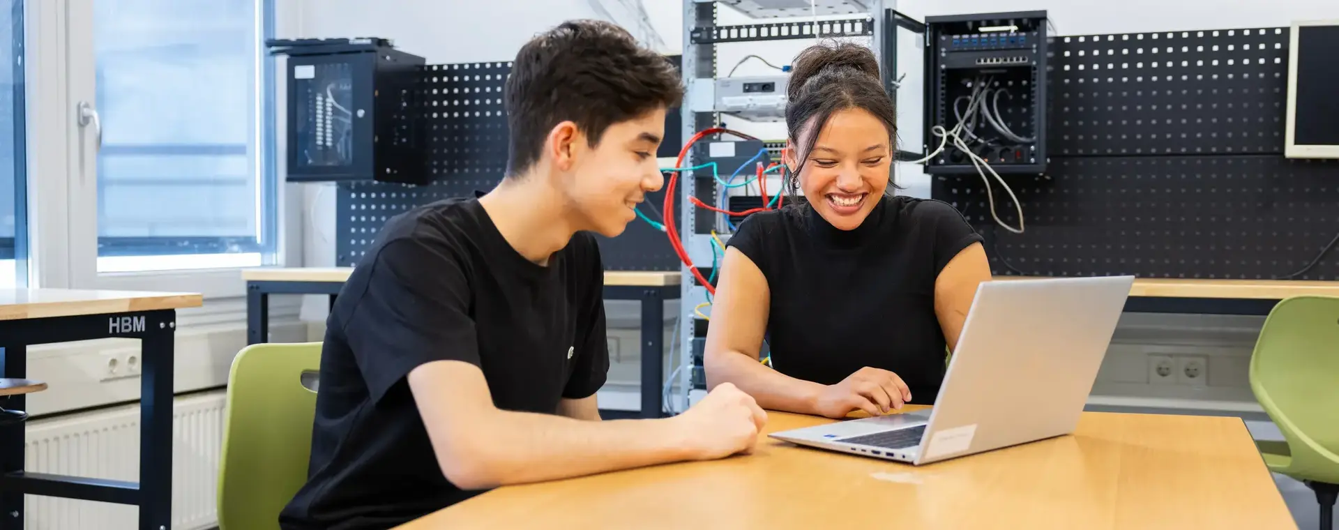 Twee studenten werken samen achter een laptop in een ICT-lokaal. Op de achtergrond staan netwerkapparaten en bekabeling opgesteld.
