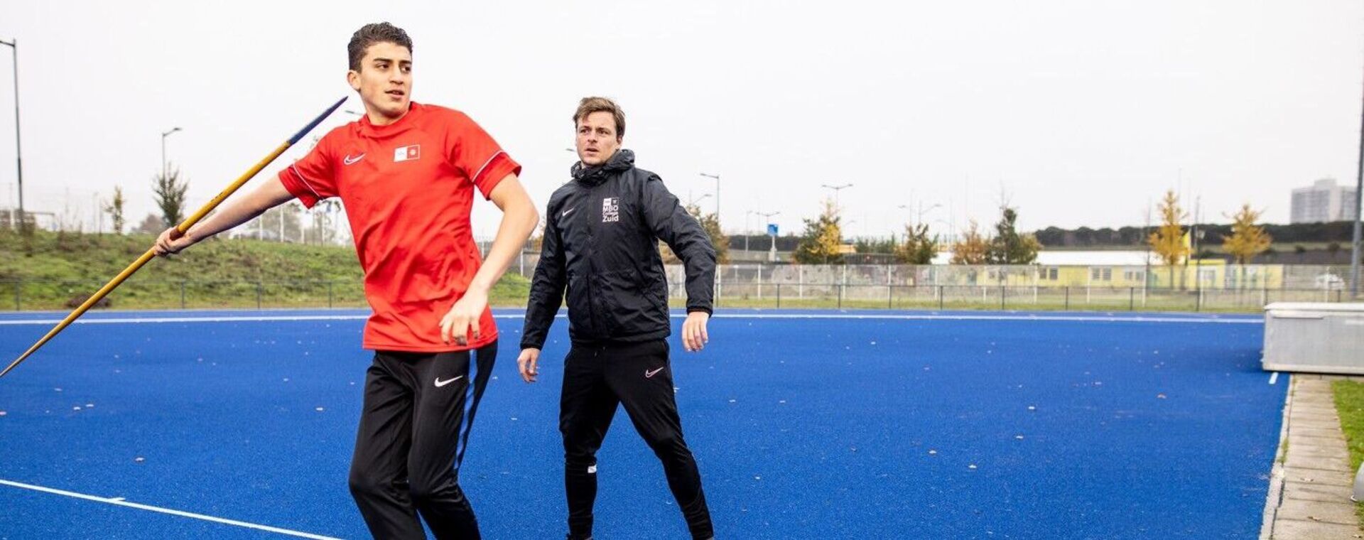 Coach kijkt toe terwijl jongen speerwerpt op atletiekbaan. 