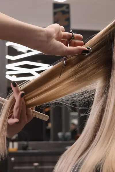 Een kapper knipt lang blond haar met een schaar en houdt het haar recht met een kam. De klant zit in een stoel in de salon.