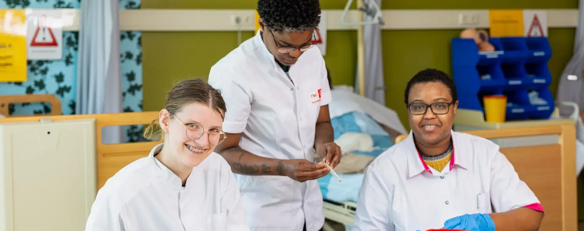 Drie studenten in witte uniformen oefenen verpleegtechnische handelingen in een zorglokaal.