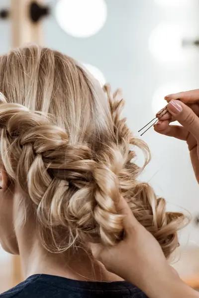 Een kapper steekt het blonde haar van een klant op in een vlecht met haarspelden. De klant zit voor een spiegel met lampen.