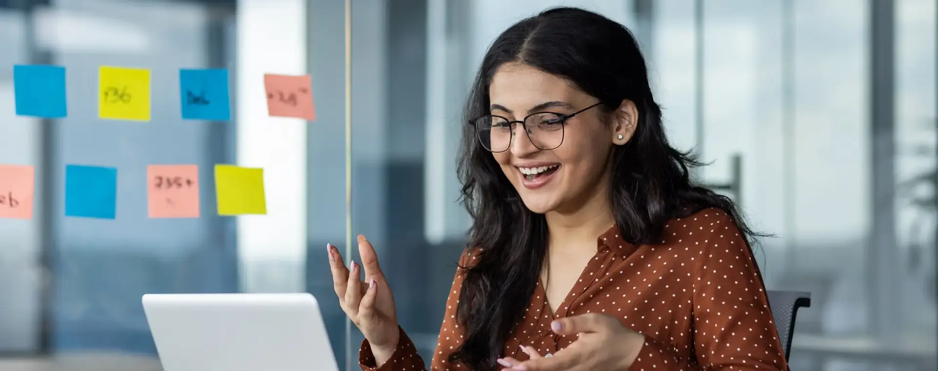 Vrouwelijke student met bril zit achter een laptop in een glazen kantoorruimte en reageert enthousiast tijdens een videogesprek. Achter haar hangen kleurrijke post-its op het raam.