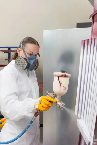 Student met een masker sprayt een coating op een paneel terwijl een docent met een masker toekijkt en aanwijzingen geeft.
