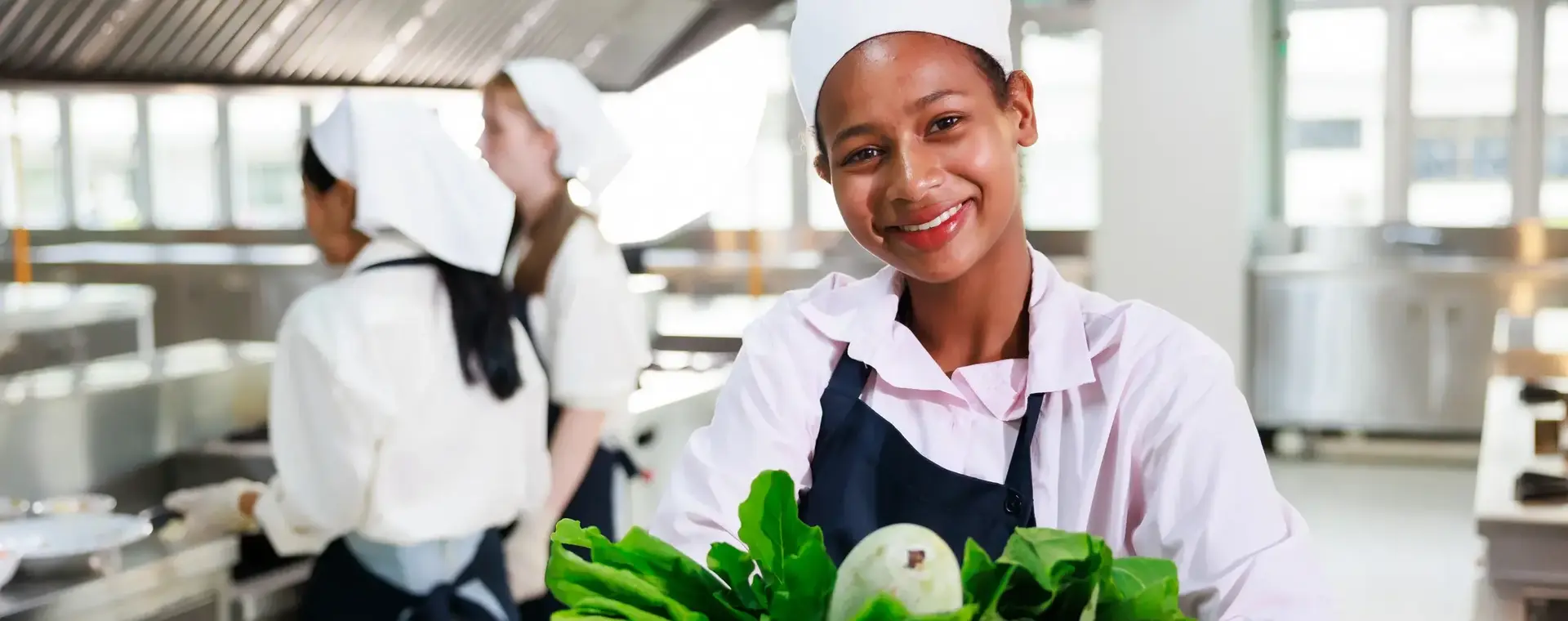 Student in opleiding toont verse groenten in keuken met twee koks op de achtergrond