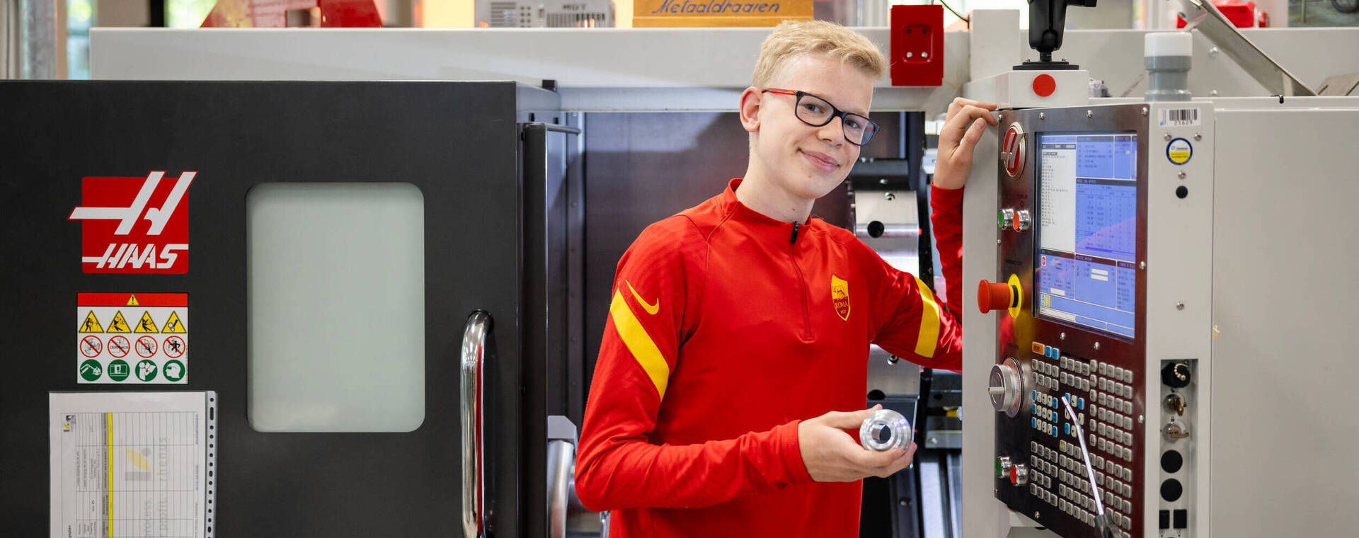 Student in rood shirt staat bij een CNC-machine en toont een metalen onderdeel dat hij heeft gemaakt.