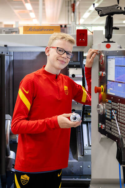 Student in rood shirt staat bij een CNC-machine en toont een metalen onderdeel dat hij heeft gemaakt.
