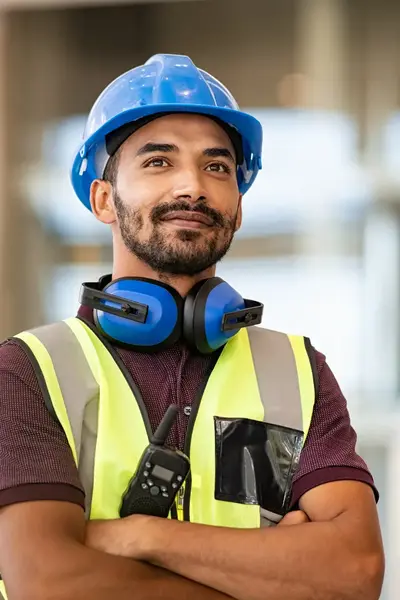 Een man met helm, gehoorbescherming en een geel hesje staat met zijn armen over elkaar en glimlacht een beetje. Op de achtergrond zie je een gebouw.