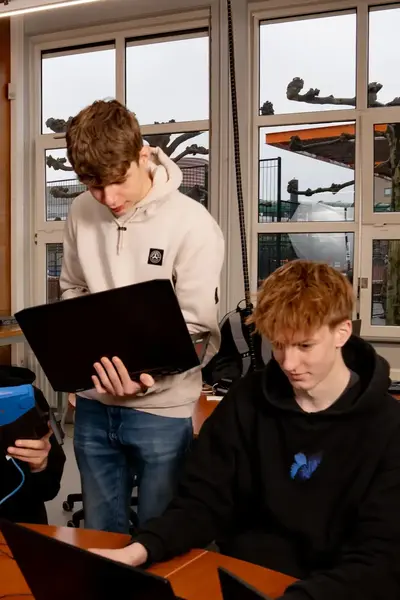 Vier studenten werken samen in een klaslokaal aan laptops en IT-apparatuur, terwijl één student een netwerkapparaat bekijkt.