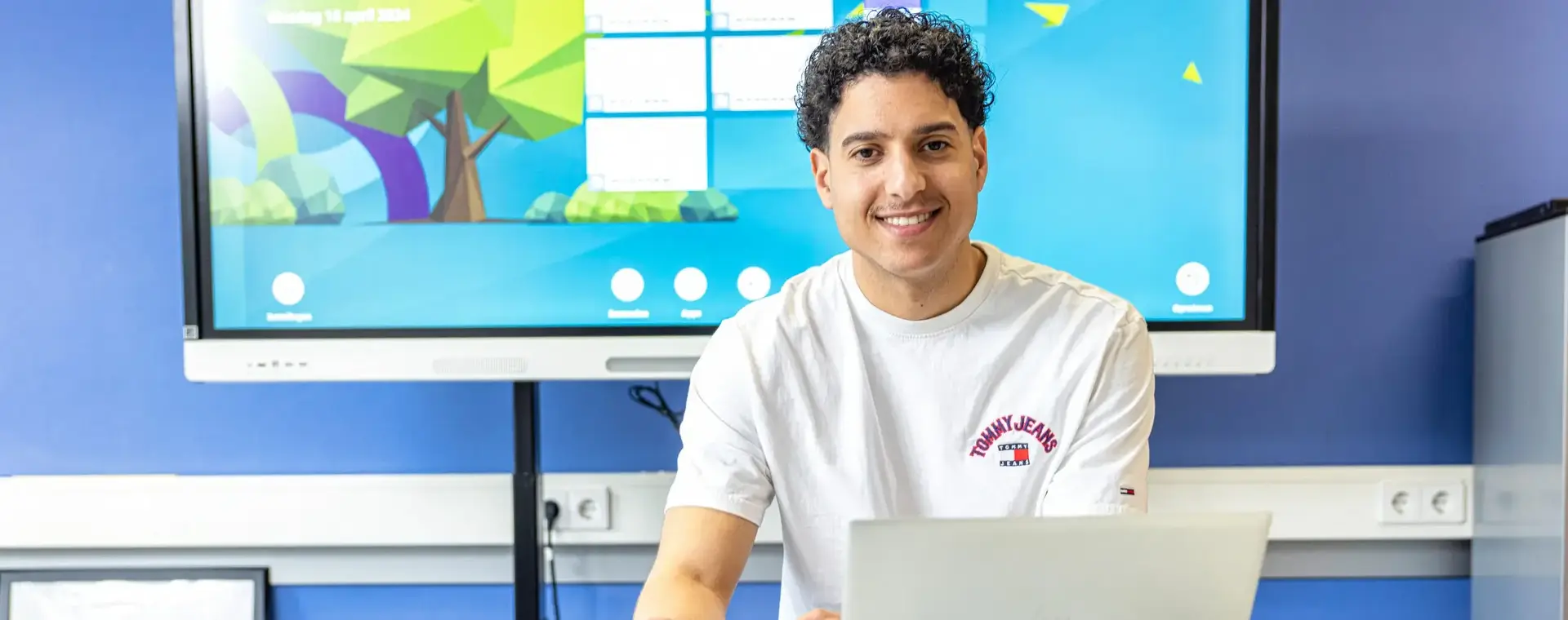 Docent zit achter een laptop in een klaslokaal en kijkt lachend naar de camera.