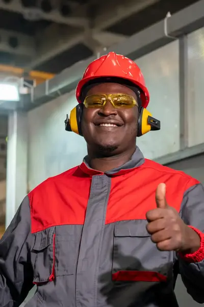 Student met ROCvA-overall, veiligheidsbril en oorbeschermers poseert naast machine in fabriek en steekt zijn duim op.