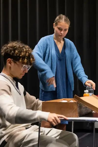 Drie studenten spelen een scène in een zwarte theaterzaal met een kist en tafel als decorstukken.