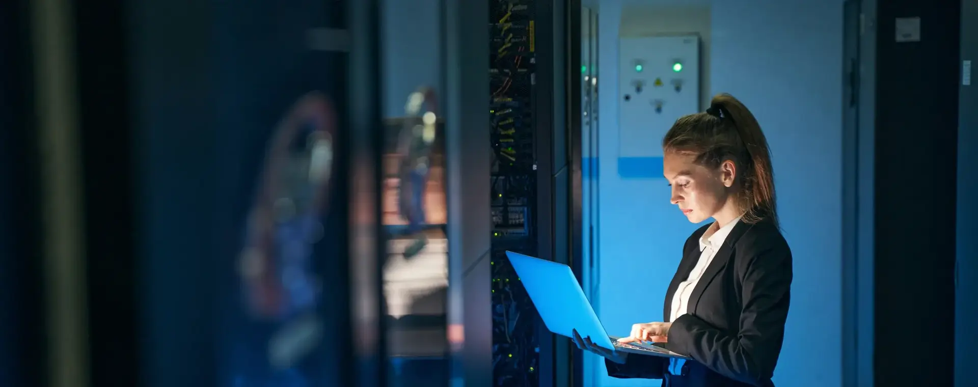 Student werkt met een laptop in een serverruimte. Ze draagt een donker pak en staat tussen verlichte serverkasten terwijl ze gegevens controleert.