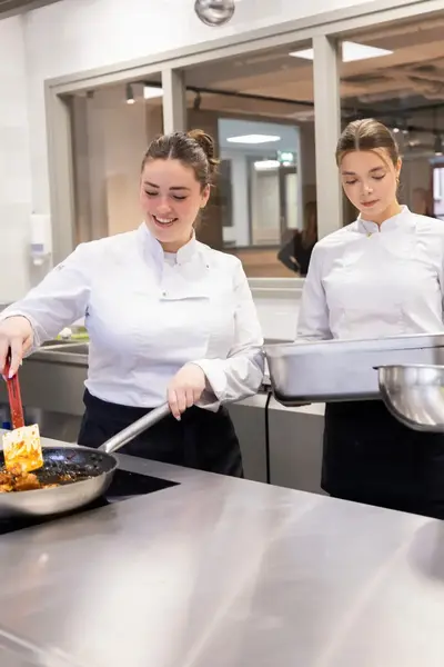 Drie studenten in kokskleding werken samen in een professionele keuken. Eén student bakt iets in een pan, terwijl de andere twee klaarstaan met een mengkom en een ovenschaal. 