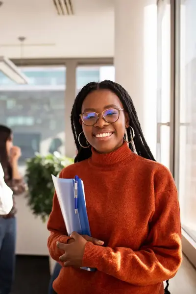 Student met bril en oranje trui staat lachend met een map in haar hand.
