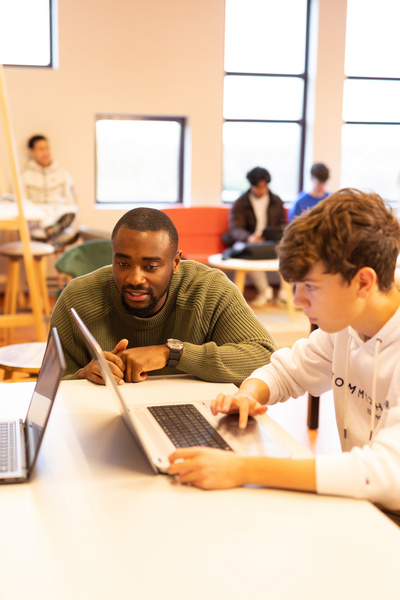 Docent begeleidt twee studenten tijdens het werken aan hun laptops in een lokaal met andere studenten op de achtergrond.