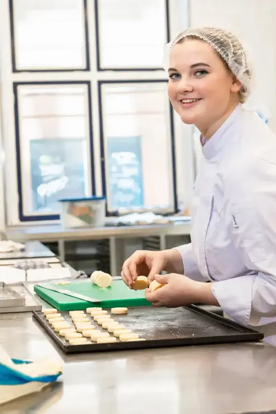 Een student in witte bakkerskleding en haarnetje staat in een bakkerij en maakt koekjes.