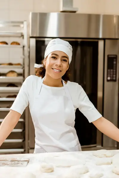 Vrouw in witte bakkerskleding staat glimlachend achter een werkblad vol deegbollen, omringd door rekken met gebakken brood.