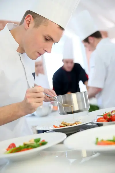 Een chef-kok in witte jas en hoge koksmuts schenkt met uiterste precisie een saus over een gerecht.