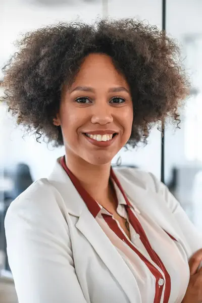 Vrouw in een lichte blazer glimlacht en staat met gekruiste armen in een kantoorruimte.