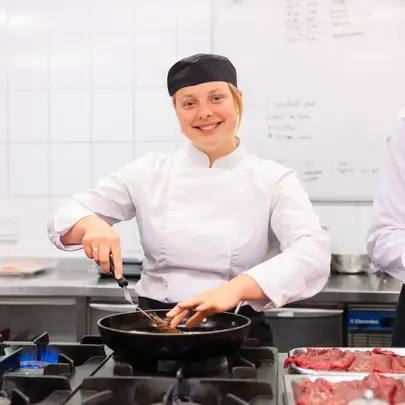 Een kok in witte jas en zwarte muts staat glimlachend te bakken in een koekenpan.