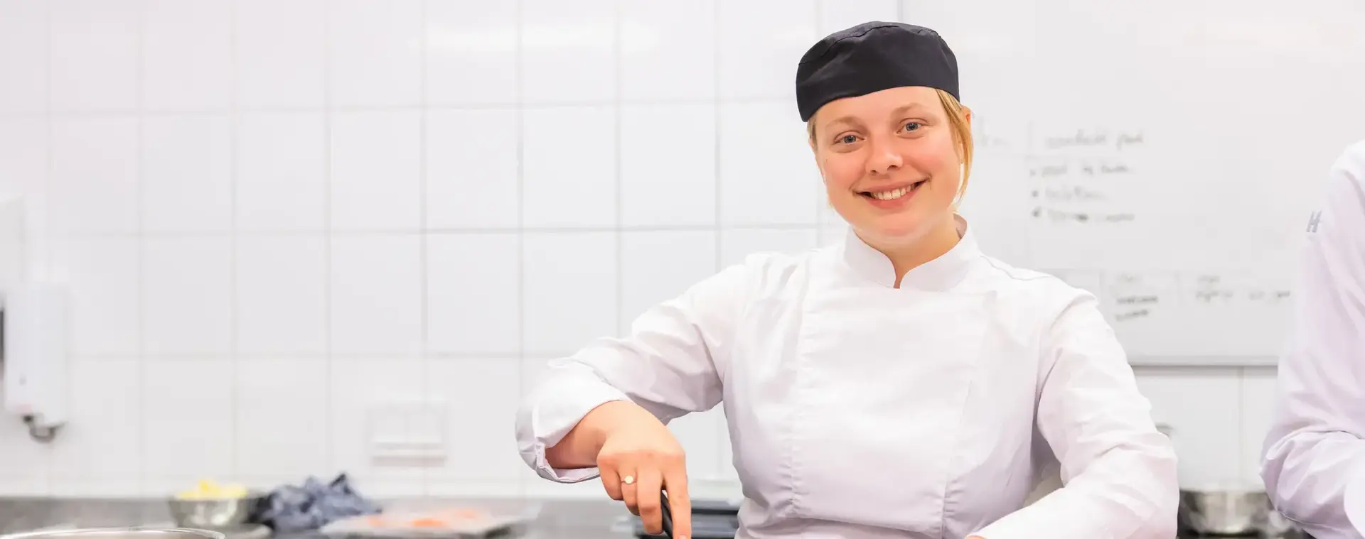 Een kok in witte jas en zwarte muts staat glimlachend te bakken in een koekenpan.