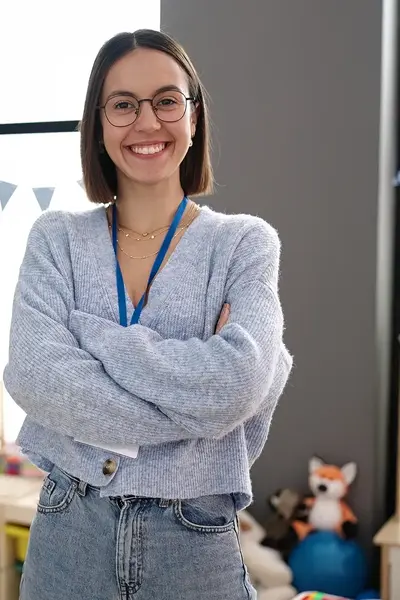 Een jonge vrouw staat lachend met haar armen over elkaar in een kleurrijke klasruimte. Op de achtergrond zijn speelgoed, knuffels en kindertekeningen te zien.