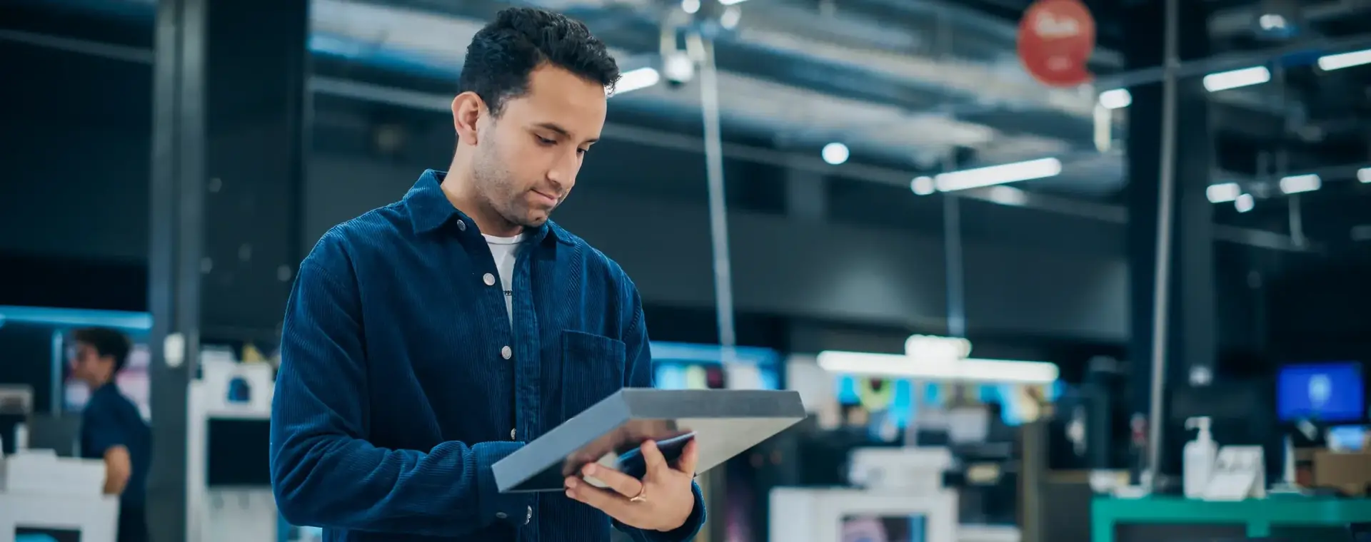 Man bekijkt een tablet in een elektronica winkel. Hij draagt een donkerblauw overhemd en staat tussen uitgestalde laptops en andere apparaten.