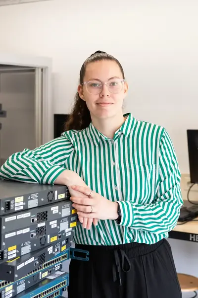 Student poseert in een ICT-lokaal naast een stapel netwerkapparatuur. Op de achtergrond zijn serverkasten en computers te zien.