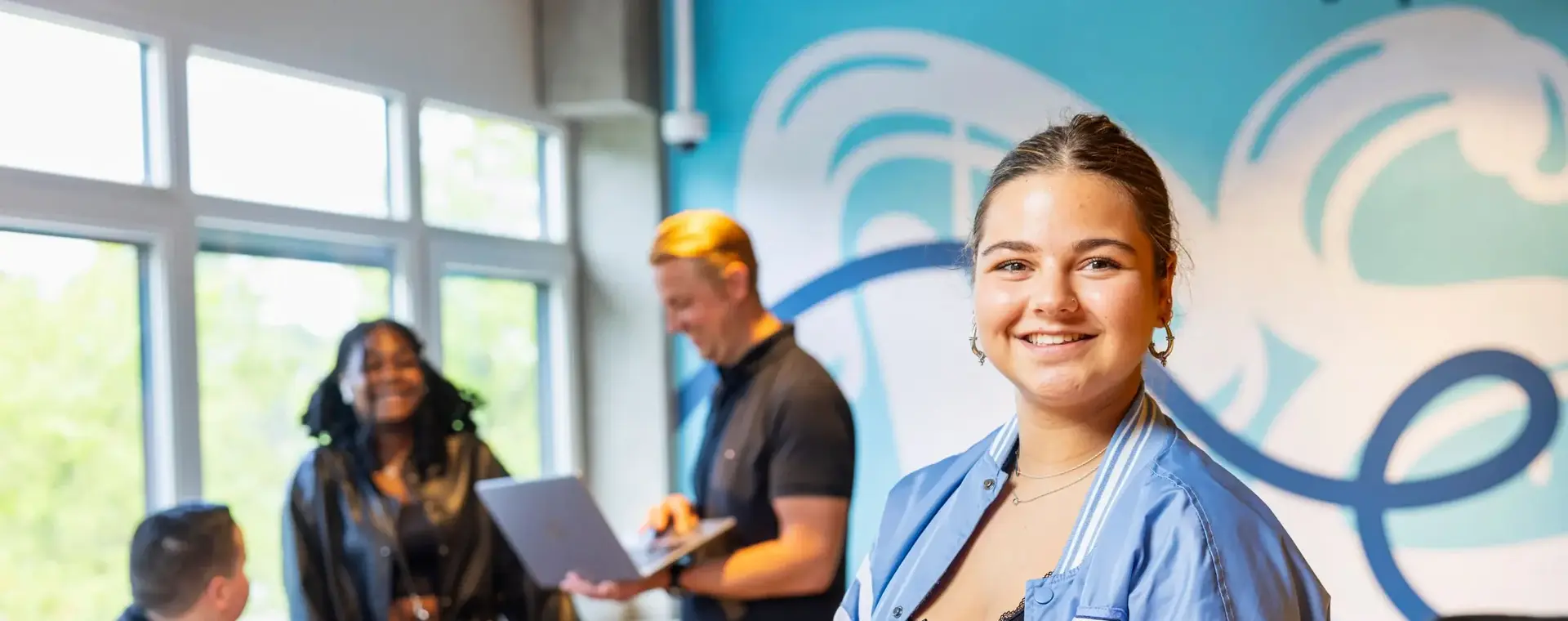 Student met laptop in haar hand lacht naar de camera, met drie andere studenten op de achtergrond,