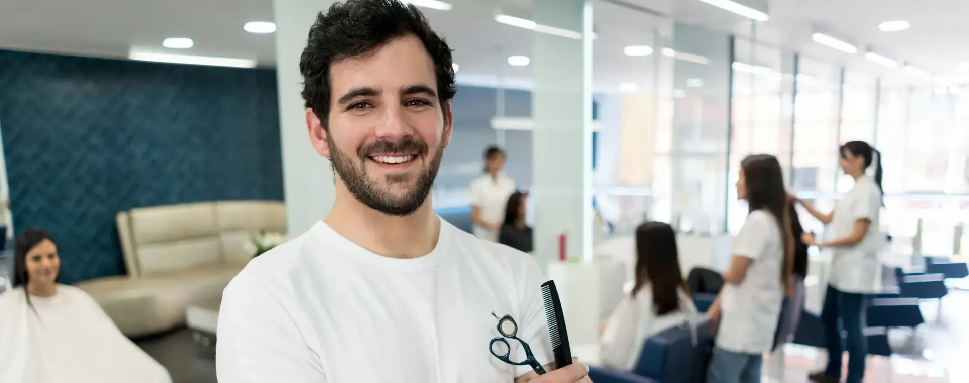Een kapper met een schaar en kam in zijn hand staat in een praktijklokaal met op de achtergrond meerdere kappersstoelen waar studenten aan het knippen zijn.