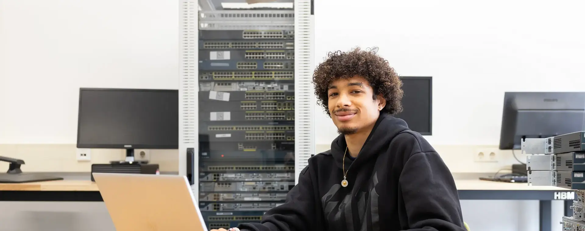 Student werkt op een laptop in een ICT-lokaal met netwerkapparatuur op de achtergrond. Hij glimlacht naar de camera terwijl hij aan tafel zit.        Ask ChatGPT