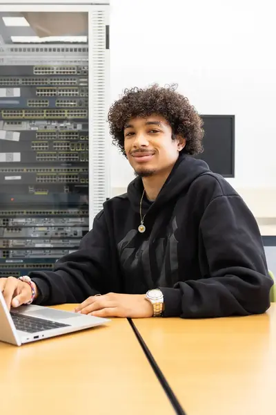 Student werkt op een laptop in een ICT-lokaal met netwerkapparatuur op de achtergrond. Hij glimlacht naar de camera terwijl hij aan tafel zit. Ask ChatGPT