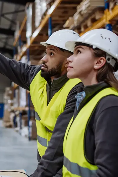 Man en vrouw in geel vestje en met helm inventariseren met behulp van de computer de voorraad.