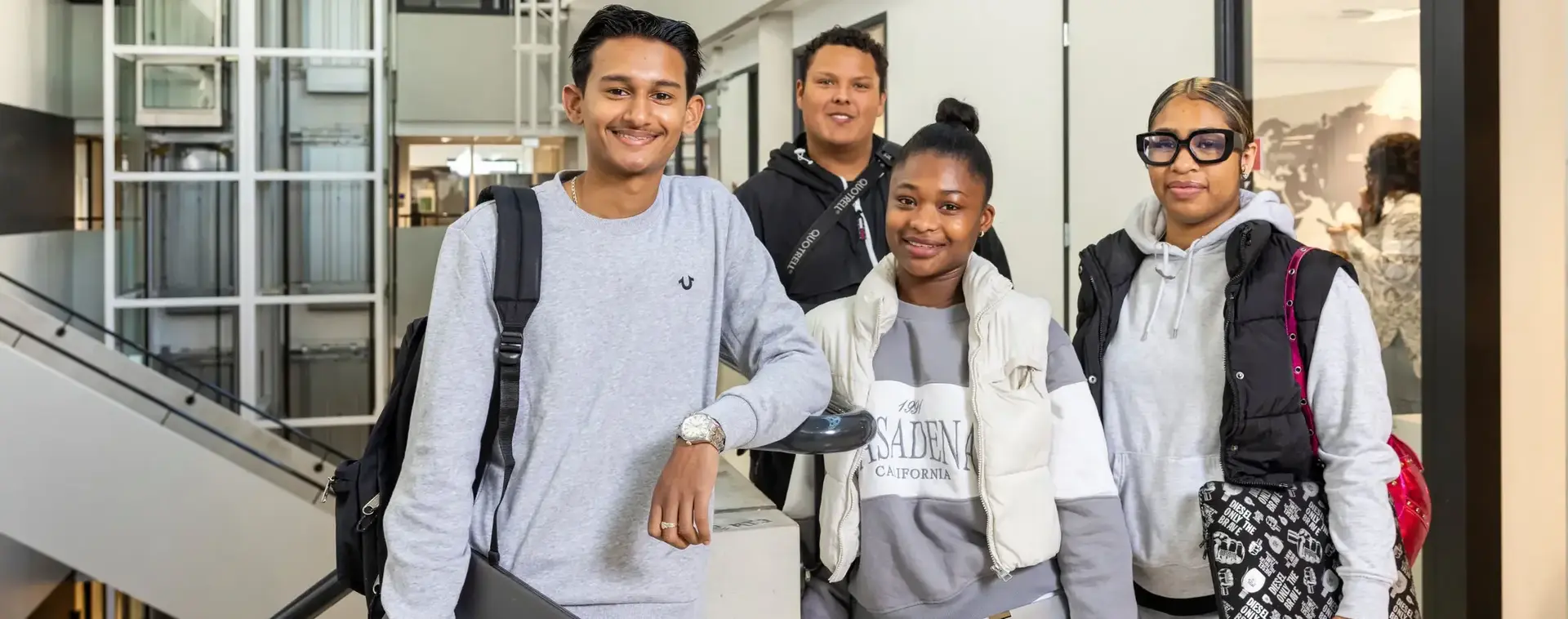 Vier studenten staan samen in de schoolgang en lachen naar de camera. Ze dragen laptops en rugzakken.