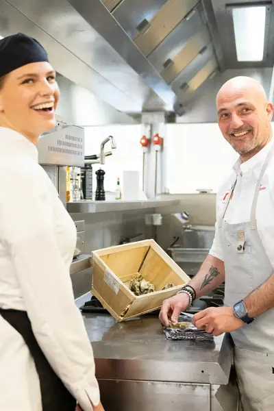 Twee koks staan lachend in de keuken. De vrouw draagt een koksmuts en de man sorteert oesters op het werkblad..