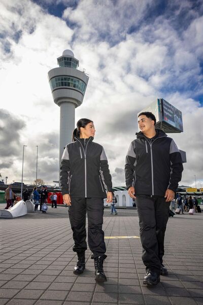 Studenten Luchthavenbeveiliger lopen op Schiphol met luchtverkeerstoren op de achtergrond